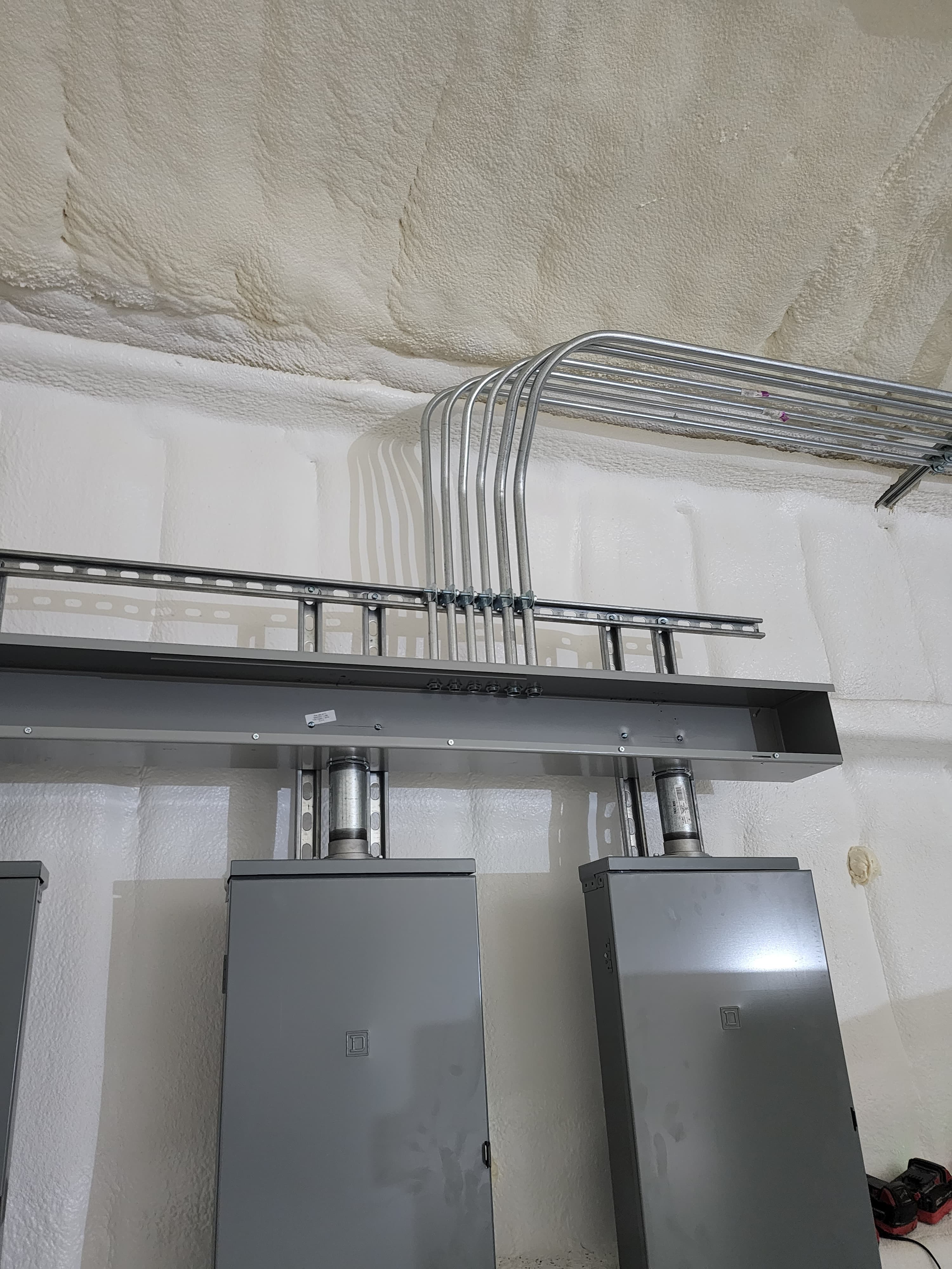 Gray electrical boxes and metal conduits mounted against a white spray foam insulated wall.