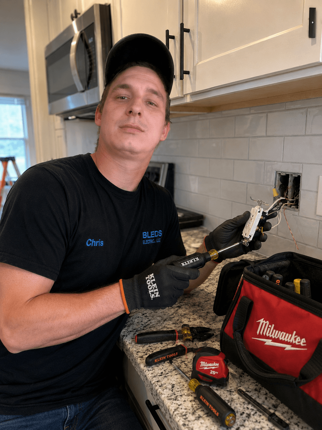 An electrician installs a white electrical outlet in a kitchen backsplash using hand tools.
