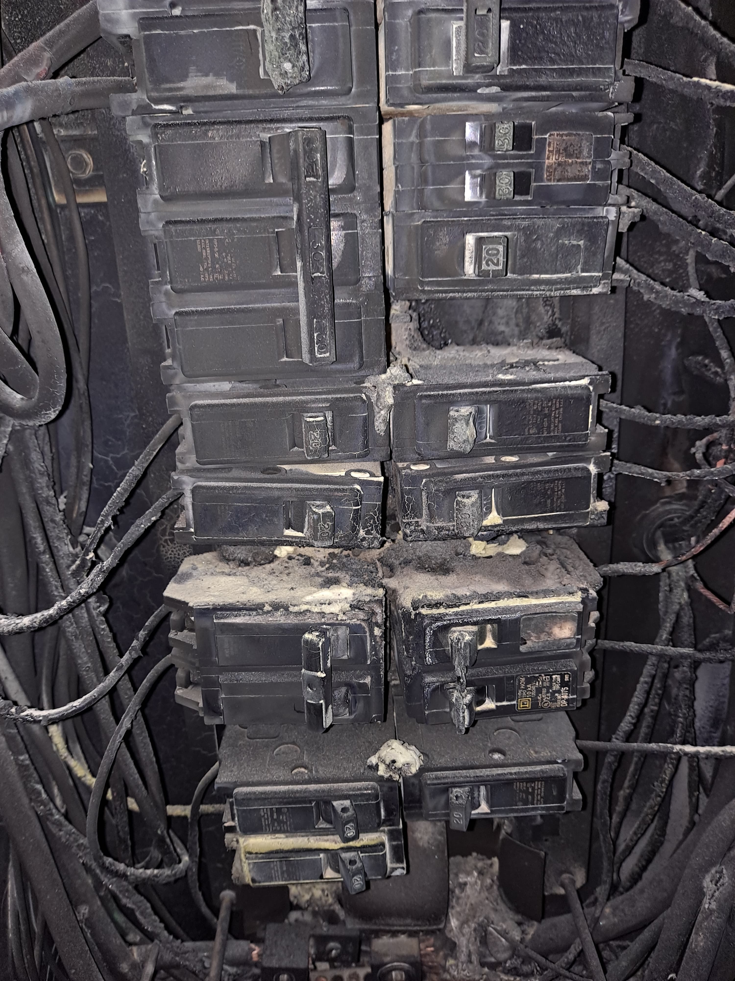 Severely charred electrical circuit breaker panel covered in black soot and melted plastic debris.