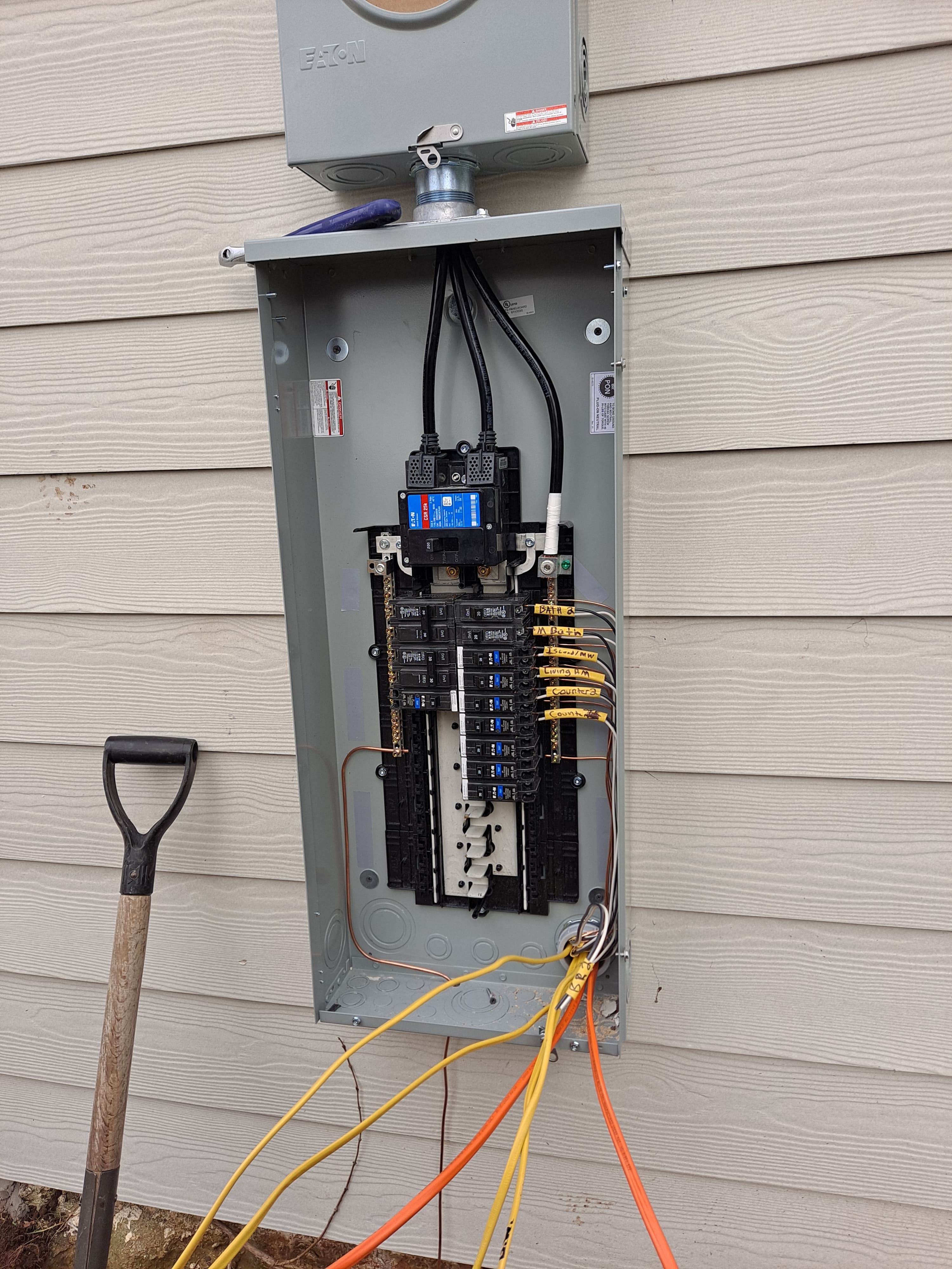 Open electrical panel on exterior siding with labeled breakers and exposed yellow and orange wires.