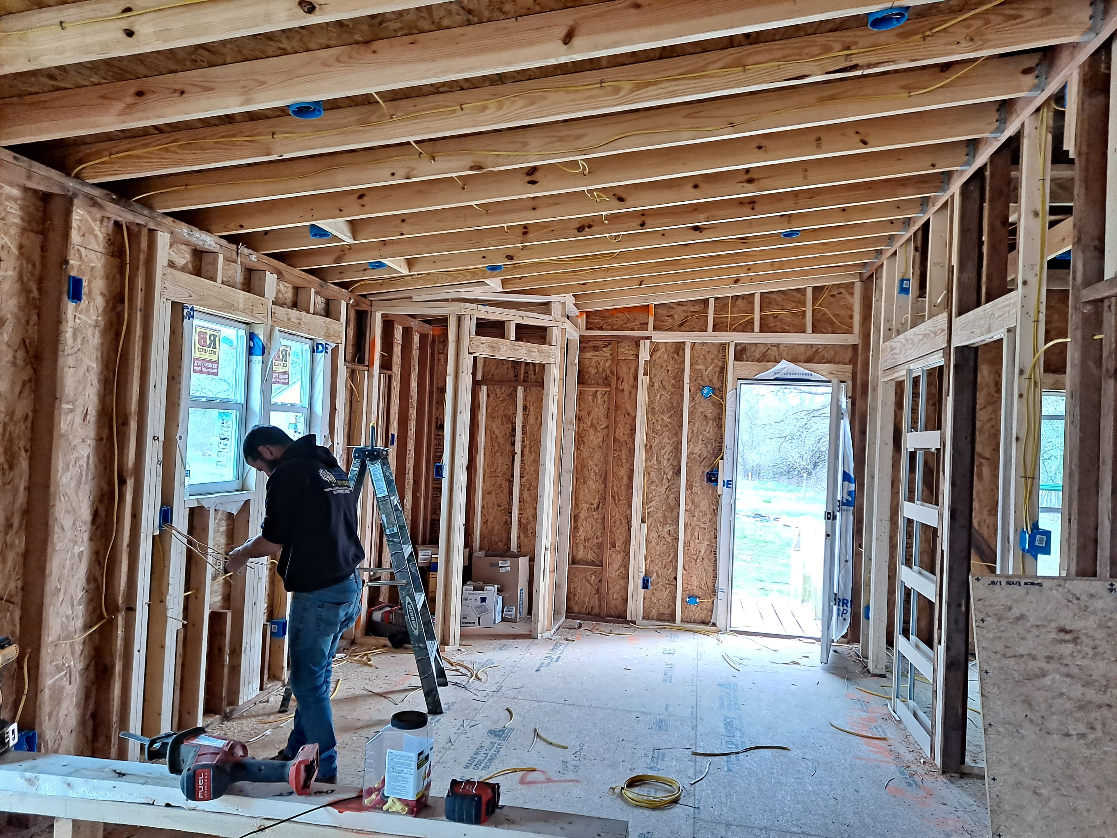 A man installs electrical wiring in a house under construction with exposed wooden framing.