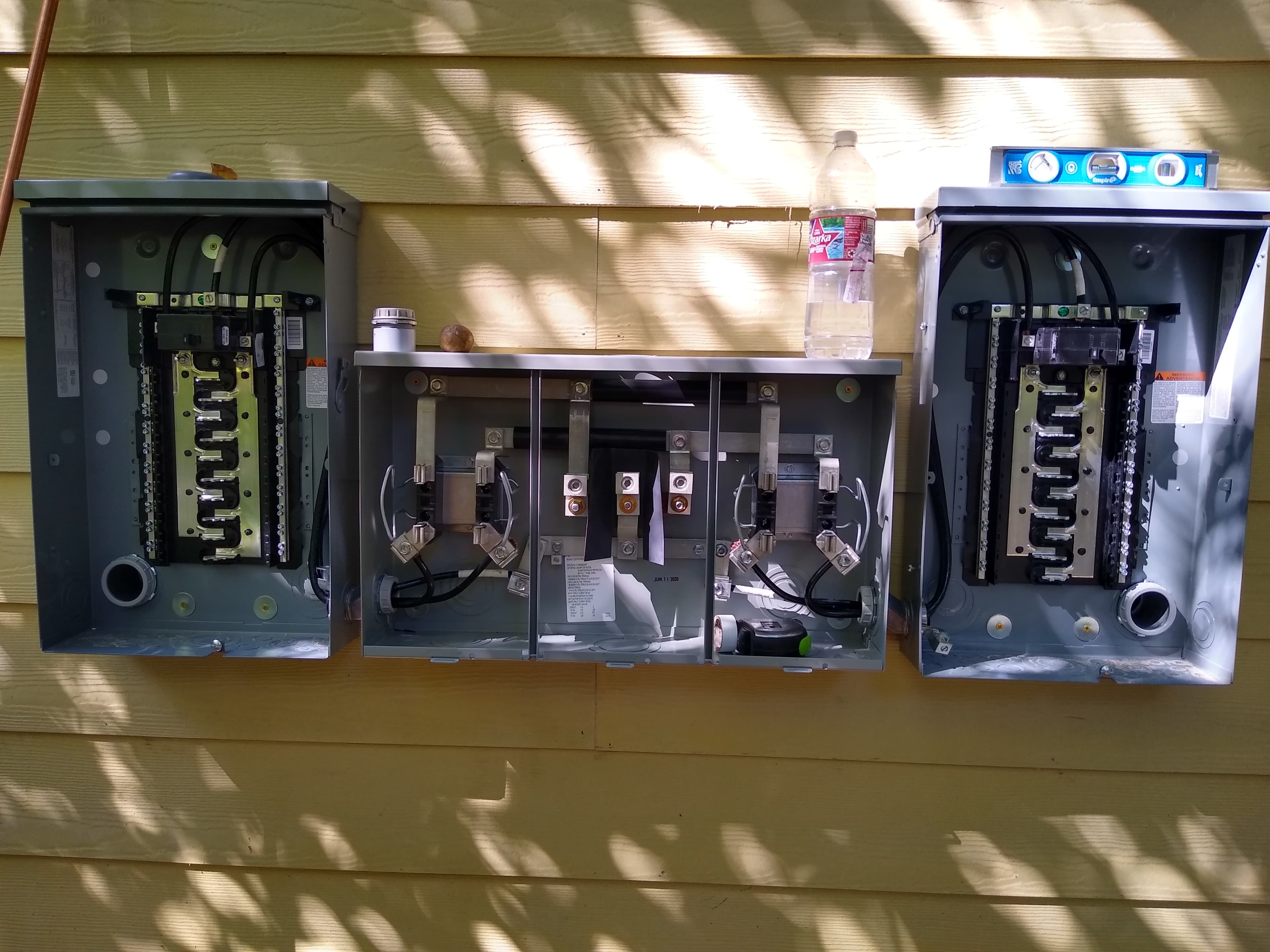 Three open electrical panels showing internal wiring mounted on yellow siding under dappled sunlight.
