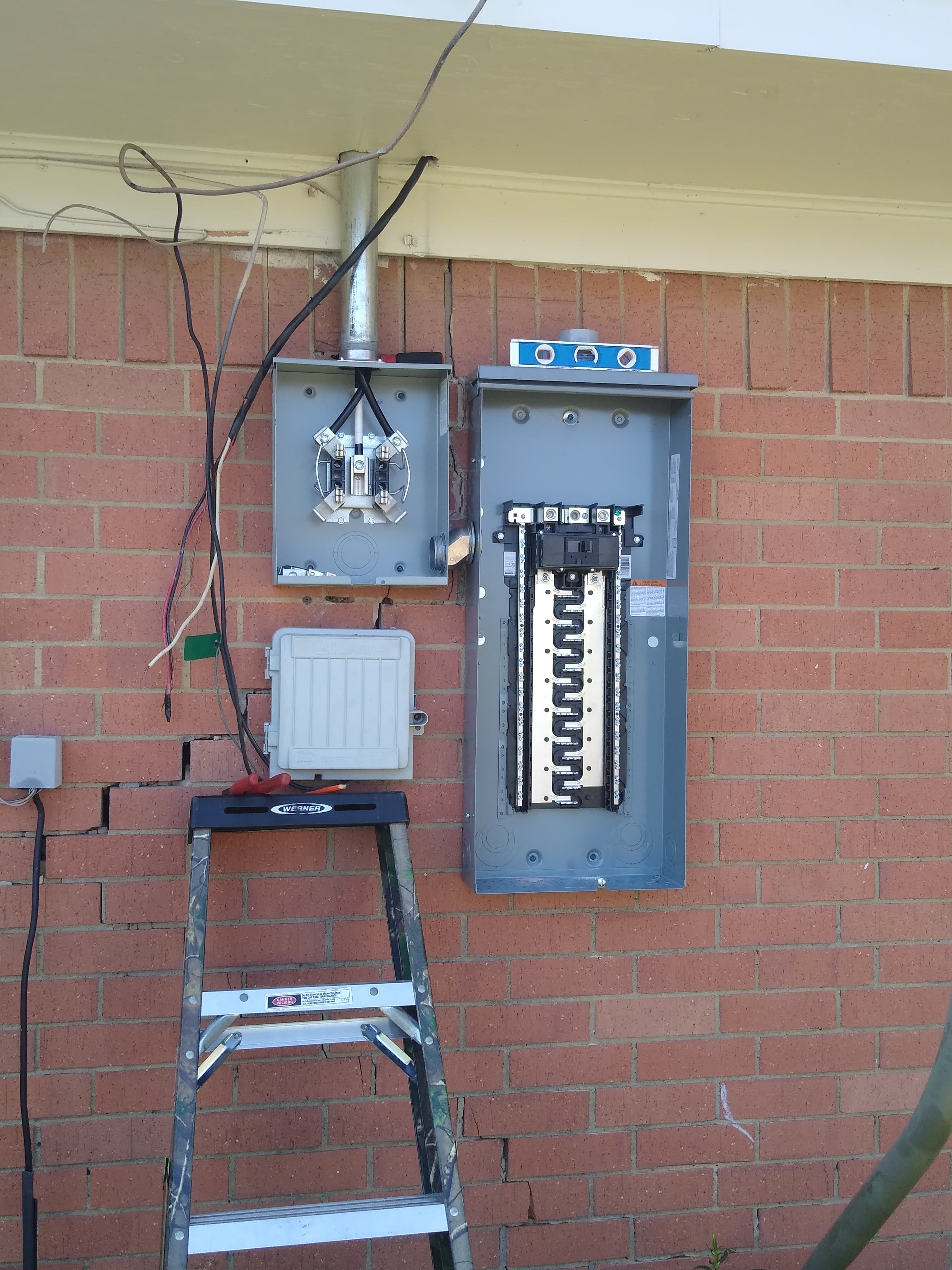 Open electrical breaker panel and meter socket being installed on a cracked red brick wall.