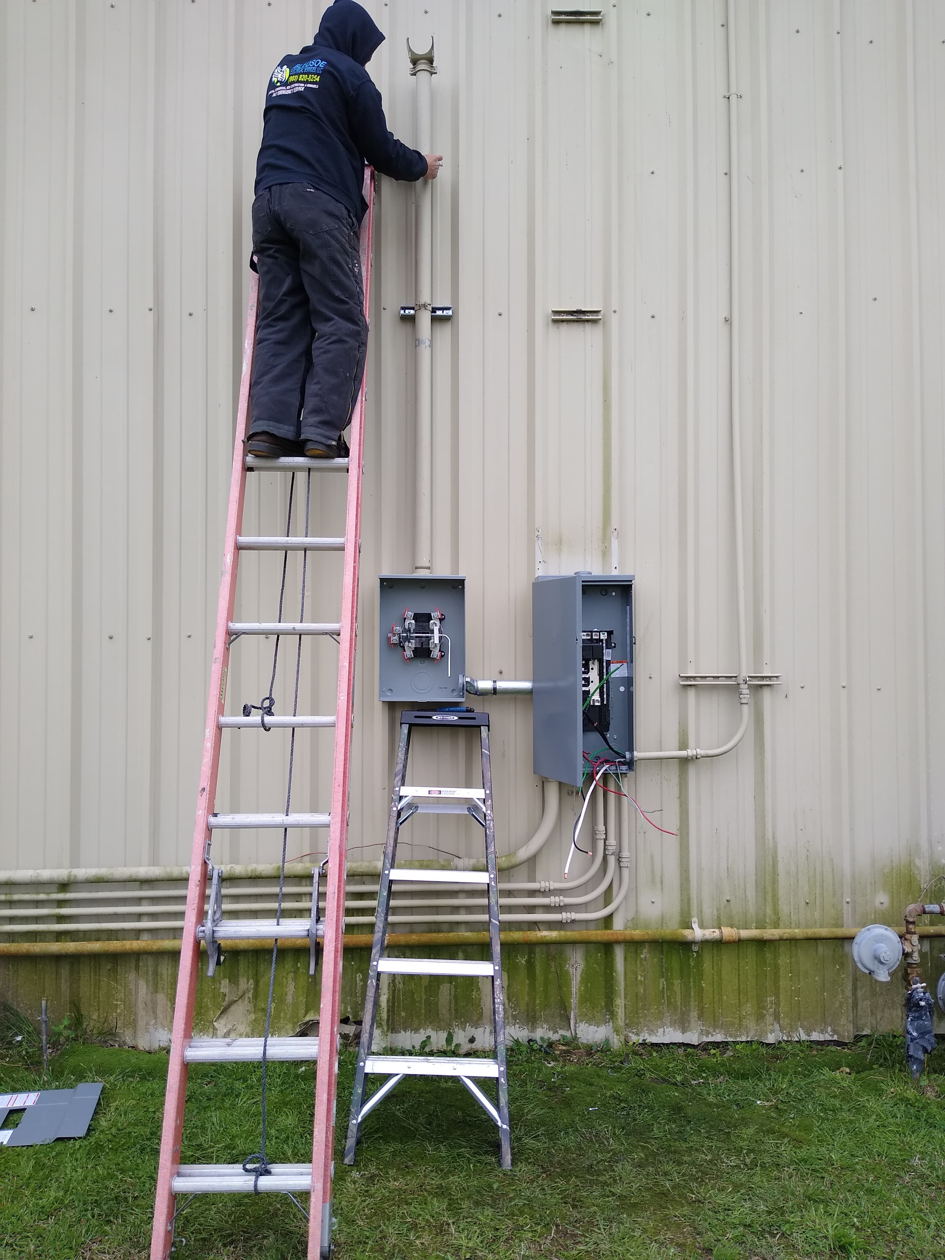 Electrician on a tall pink ladder installs electrical boxes on a beige metal wall.