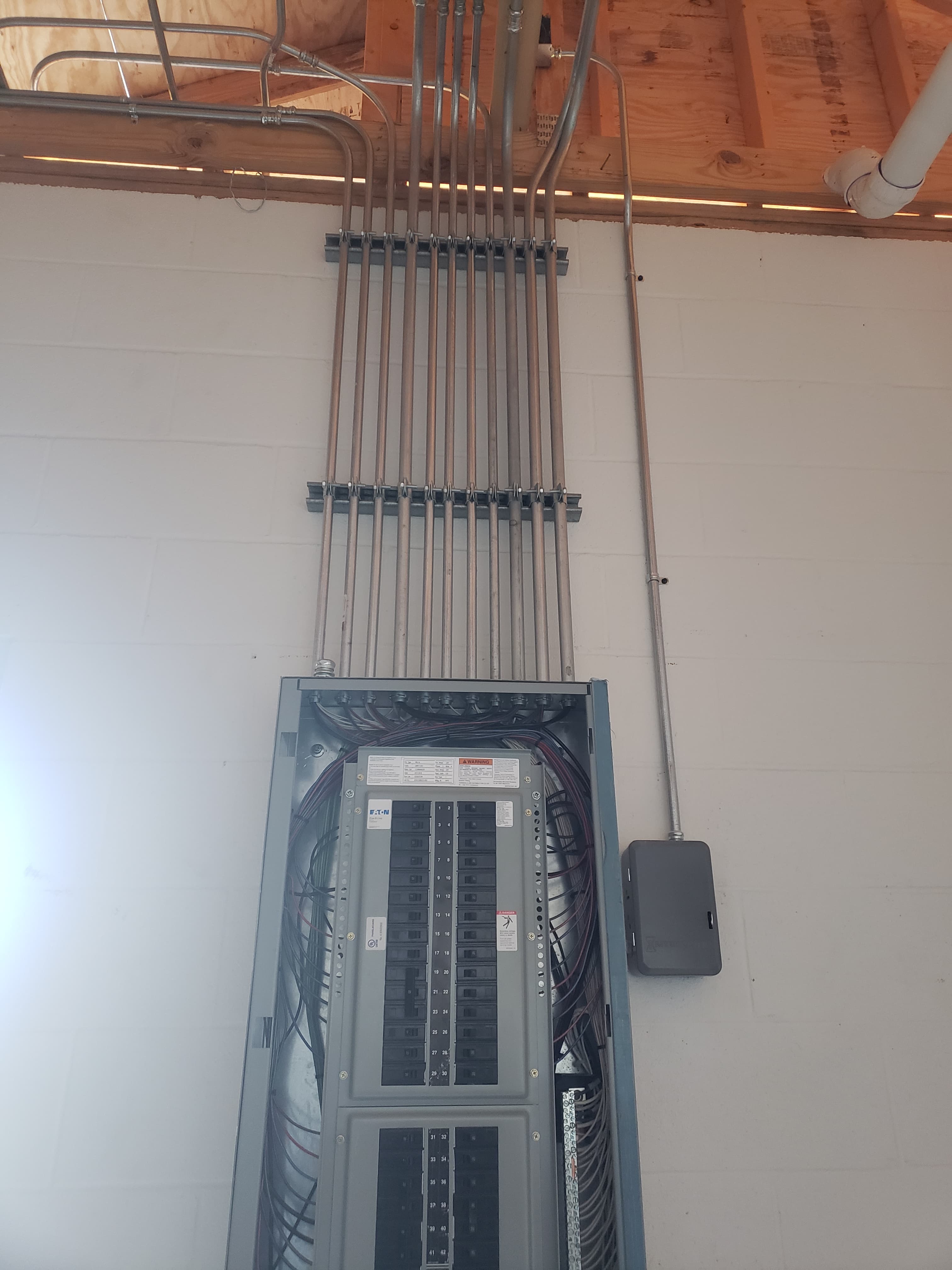 Open electrical breaker panel with vertical metal conduits mounted on a white block wall.