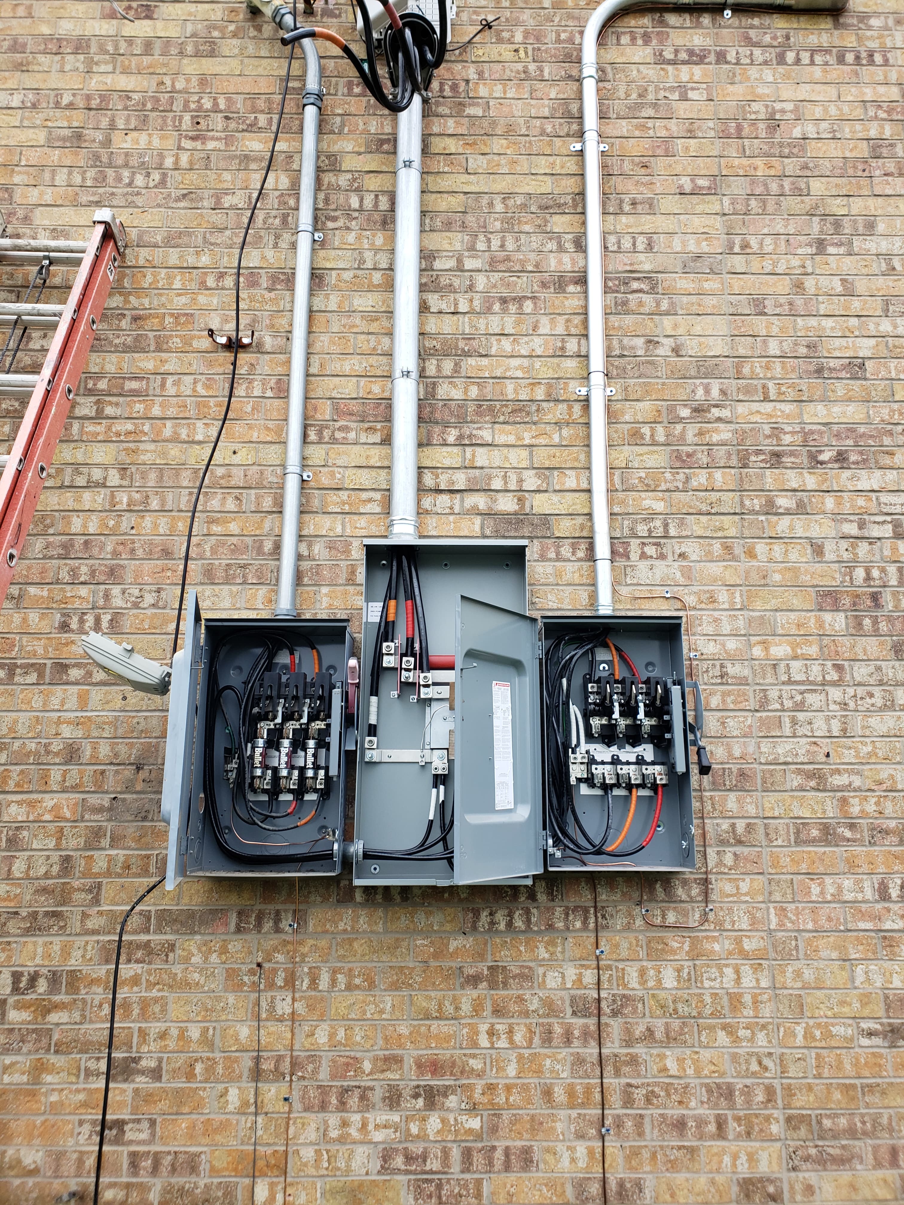 Three open electrical disconnect boxes with visible wiring and conduits mounted on a brick wall.